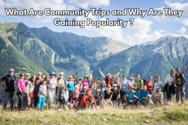 A large group of diverse travelers smiling and posing together during a mountain hike on a community trip, with scenic green mountains and blue skies in the background.